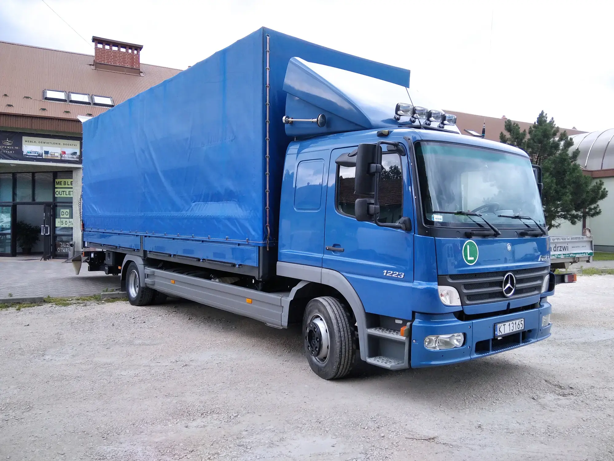 Niebieska ciężarówka Mercedes-Benz Atego 1223 z plandeką, na placu przed budynkiem